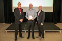 Three people pose for a photo during an awards ceremony. The person at center holds a plaque.