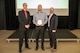 Three people pose for a photo during an awards ceremony. The person at center holds a plaque.