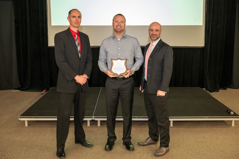 Three people pose for a photo during an awards ceremony. The person at center holds a plaque.