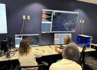 photo of military members in civilian clothing wearing headsets sitting desks with computer screens in front of them and one large screen above them