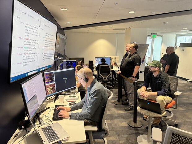 people in civilian clothes sit working on computers while others stand or sit behind them watching