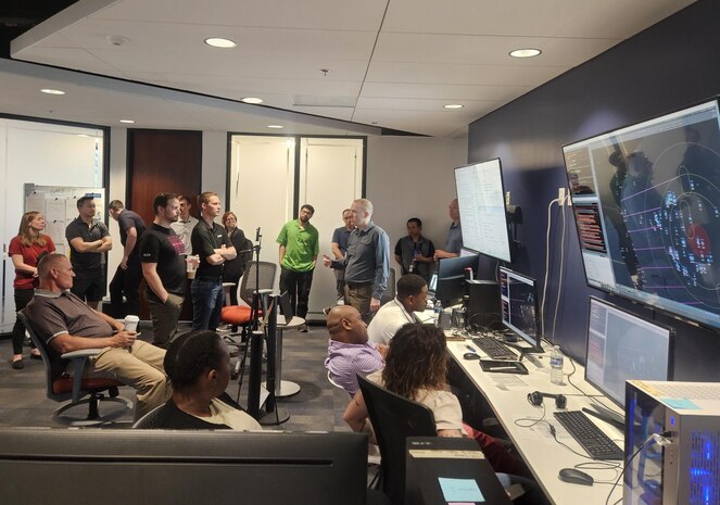 civilians sitting in chairs and standing listen to the leader standing, there are large computer screens mounted on the wall behind him and people working on computers