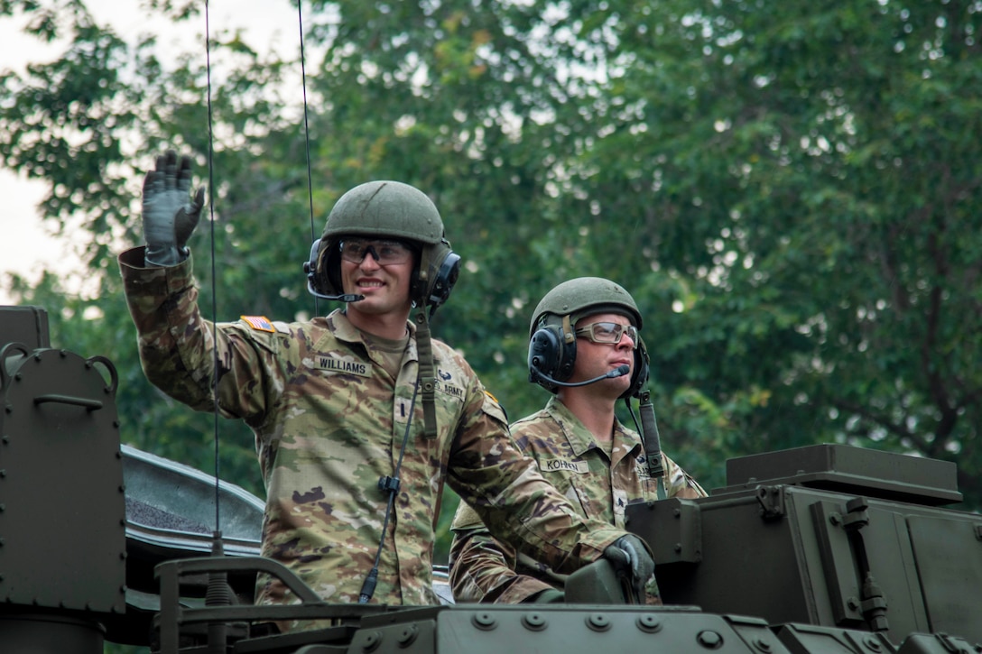 A soldier waves a gloved hand as another soldier looks forward from the perch of a vehicle with trees in the background.