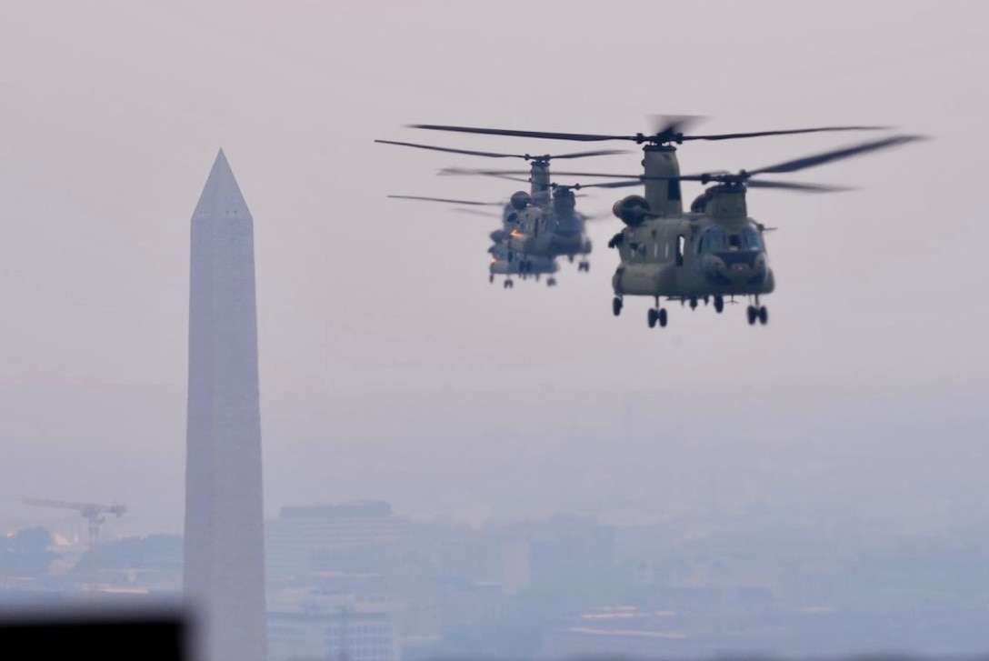 Three helicopters fly on a hazy day with a monument and buildings in the background.