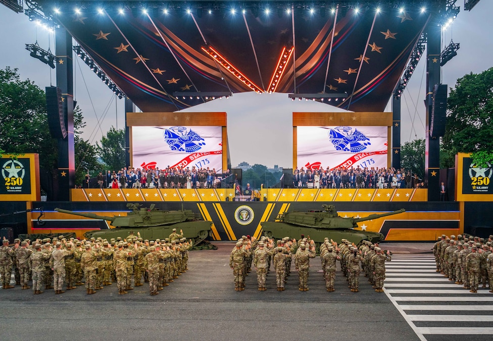 Soldiers stand with right hands raised in front of an outdoor stage; two tanks are in front of the stage, where people are seated.