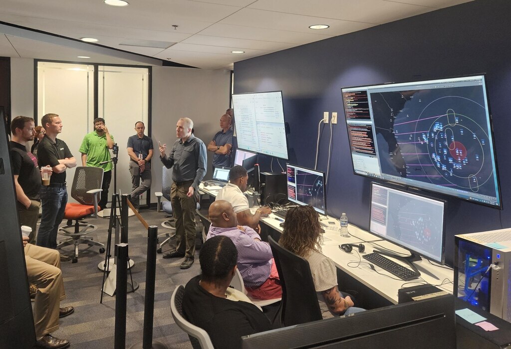 civilians sitting in chairs and standing listen to the leader standing, there are large computer screens mounted on the wall behind him and people working on computers