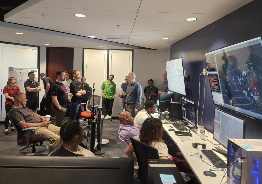 civilians sitting in chairs and standing listen to the leader standing, there are large computer screens mounted on the wall behind him and people working on computers