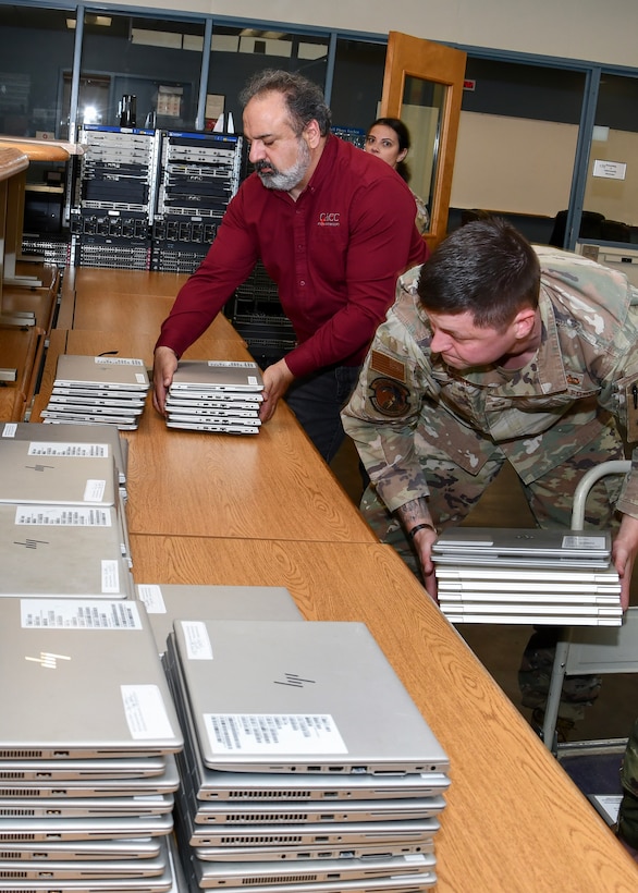 A man in a red shirt stacks laptops on a table. there is two people in air force uniforms on either side of him doing the same.