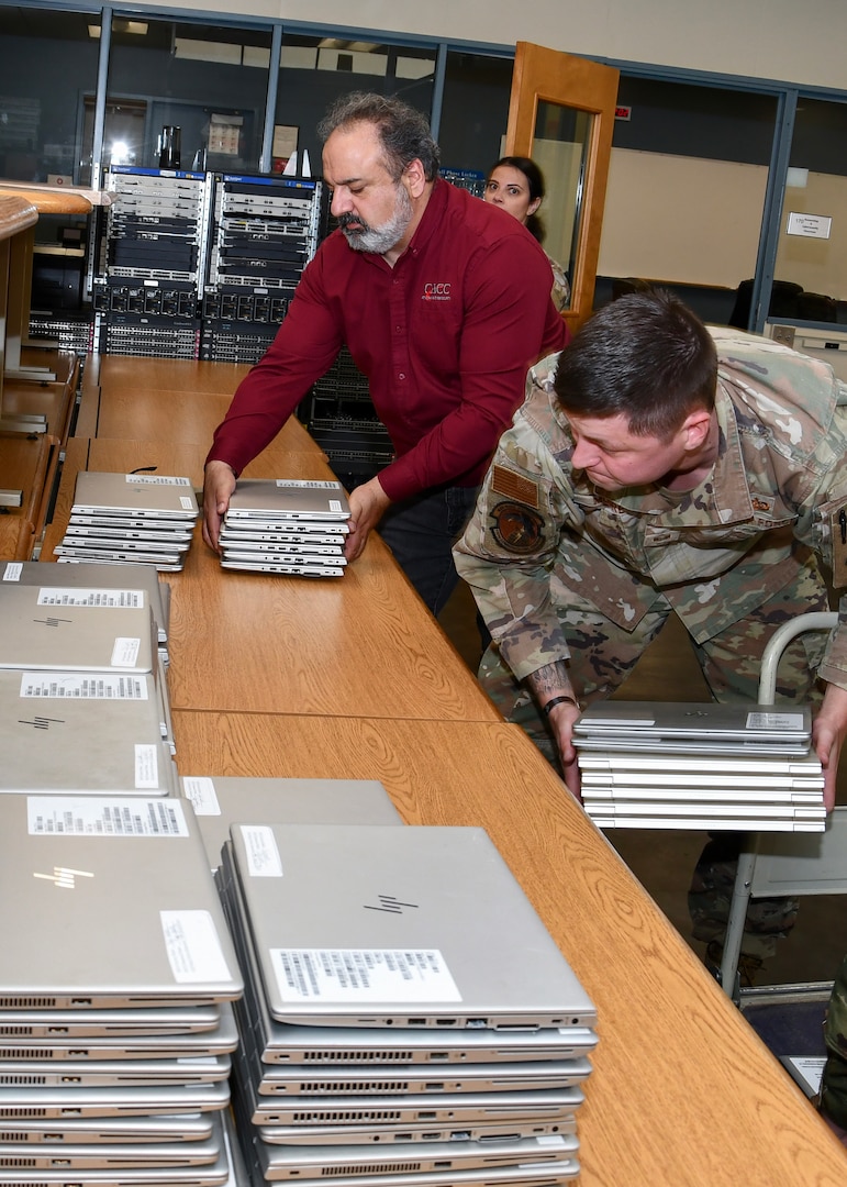 A man in a red shirt stacks laptops on a table. there is two people in air force uniforms on either side of him doing the same.