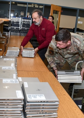 A man in a red shirt stacks laptops on a table. there is two people in air force uniforms on either side of him doing the same.