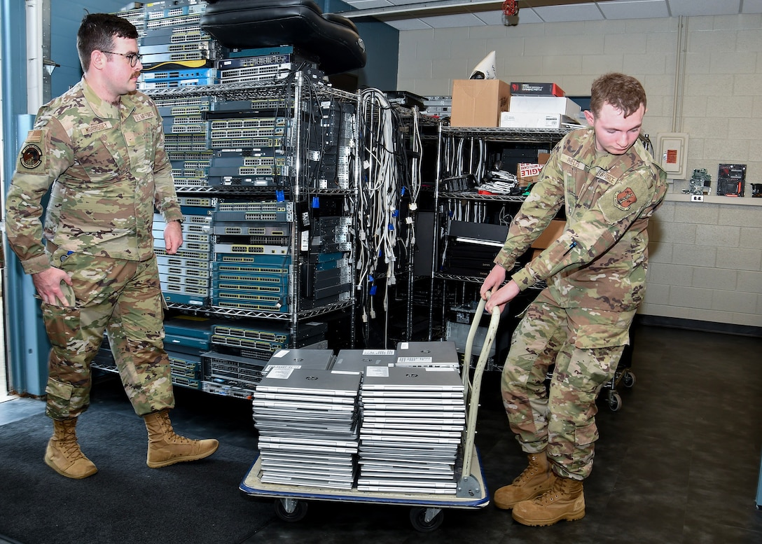Two men in Air Force Camo uniforms, one pulling a cart with a large stack of laptops the other following behind.
