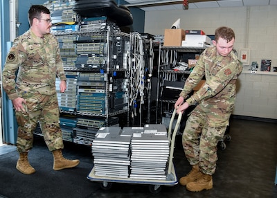 Two men in Air Force Camo uniforms, one pulling a cart with a large stack of laptops the other following behind.