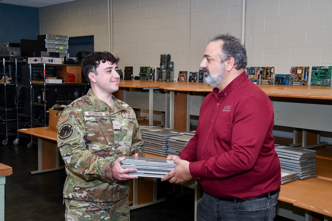 A man un an Air Force camo uniform hands a man in a red shirt a short stack of laptops.