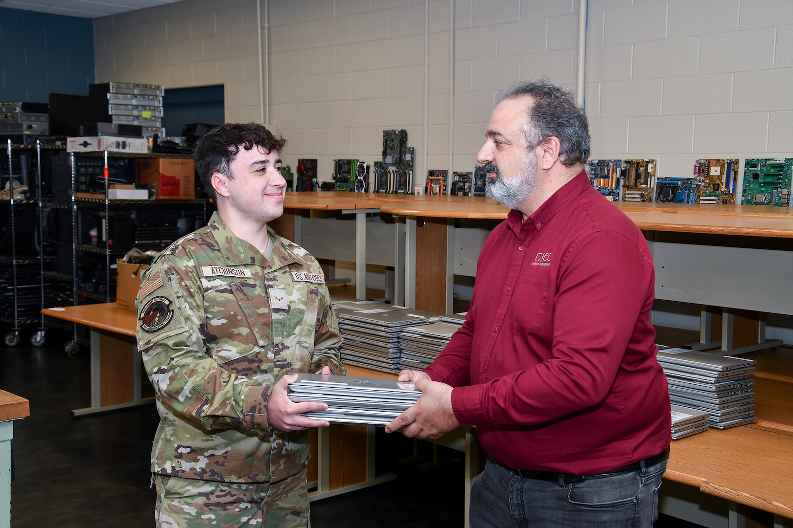 A man un an Air Force camo uniform hands a man in a red shirt a short stack of laptops.