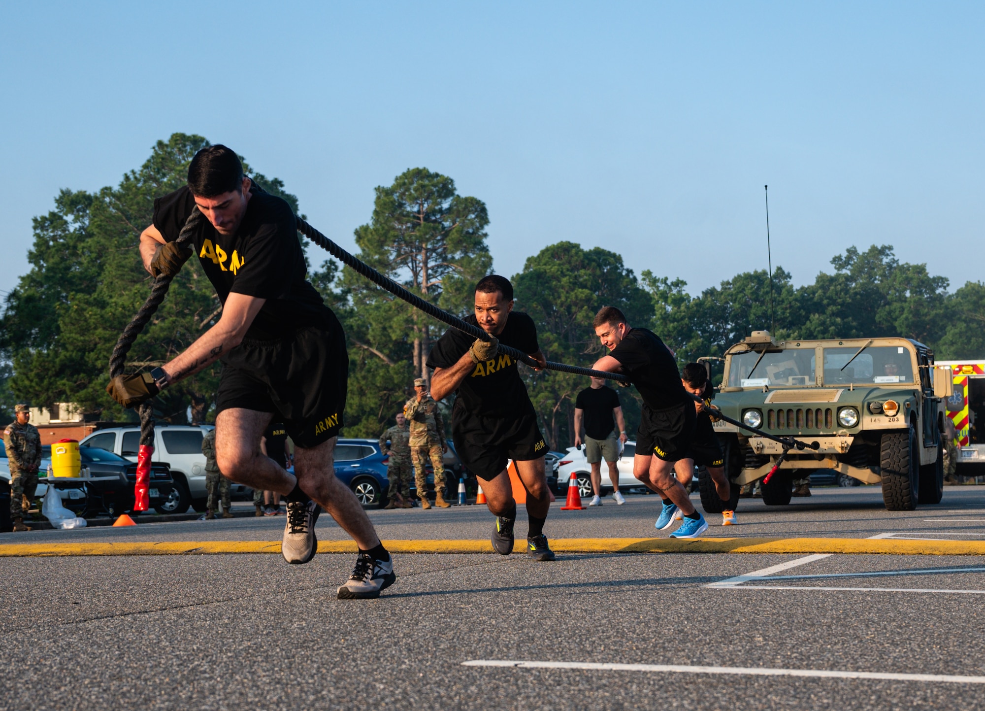 Soldiers in black pt gear pull on a rope in a line to pull a humvee outside on pavement