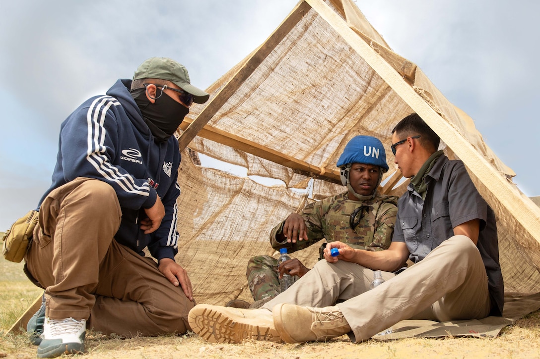 A U.S. soldier sits and talks with another person in a small tent and a third kneeling next to it in a field.