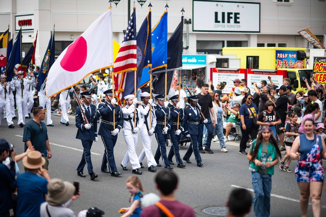 Sailors and airmen walk in formation carrying flags from different countries in a parade surrounded by dozens of spectators.