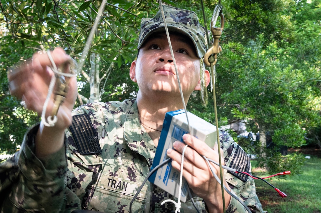 A close-up of a sailor holding the rope attached to a mosquito trap surrounded by a wooded area.