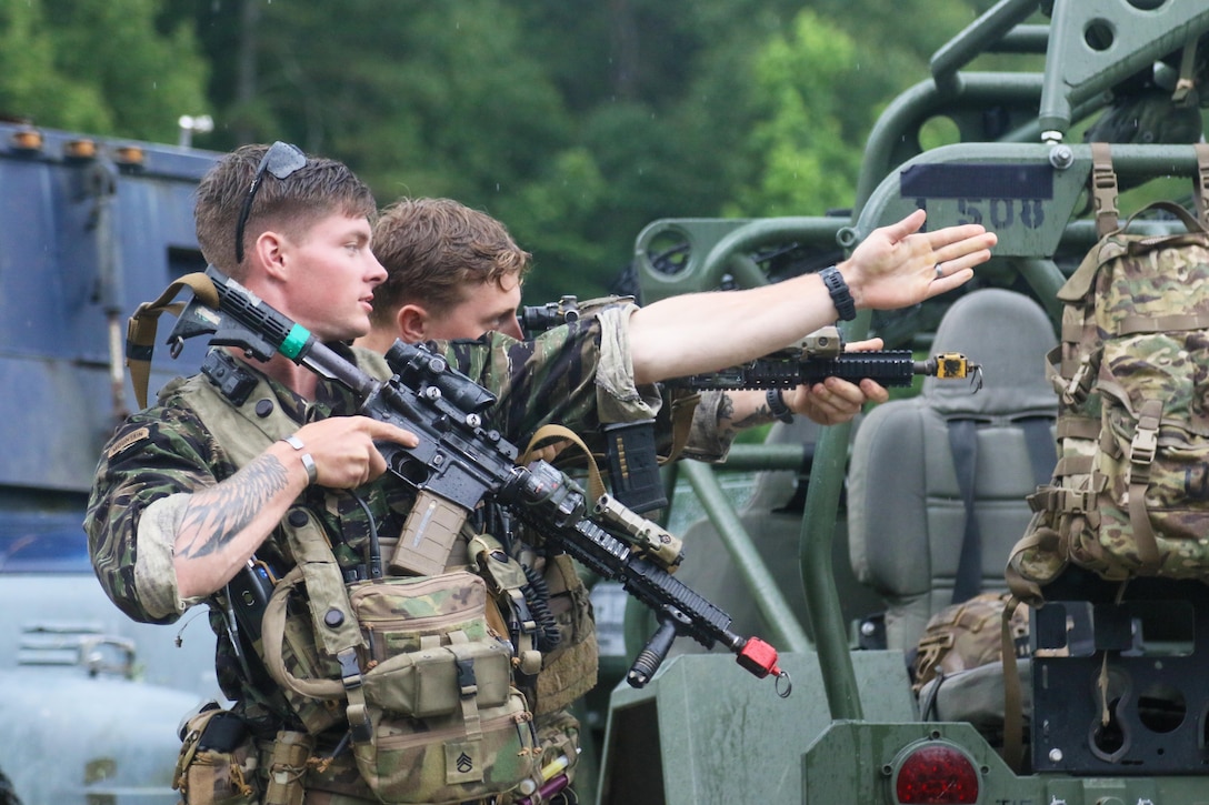 A soldier holds a weapon while pointing their arm as a fellow soldier looks through the scope of a weapon surrounded by vehicles.