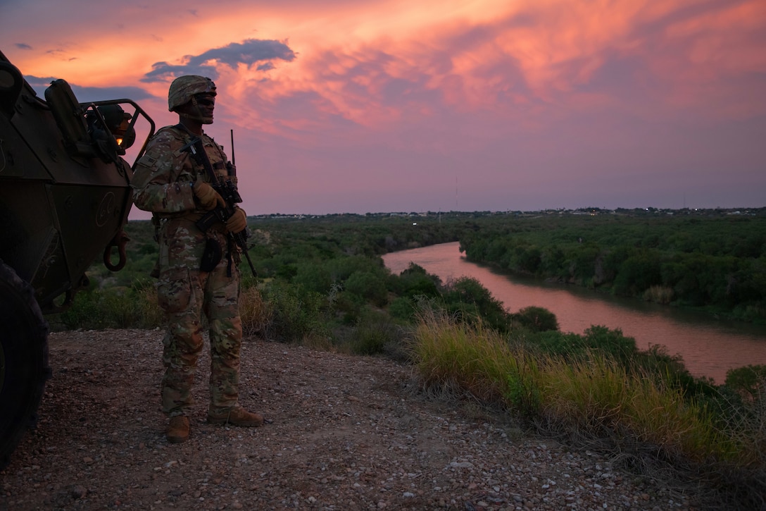 A soldier holding a rifle stands next to a vehicle parked on gravel and looks toward a river with a cloudy, orange sky in the background.