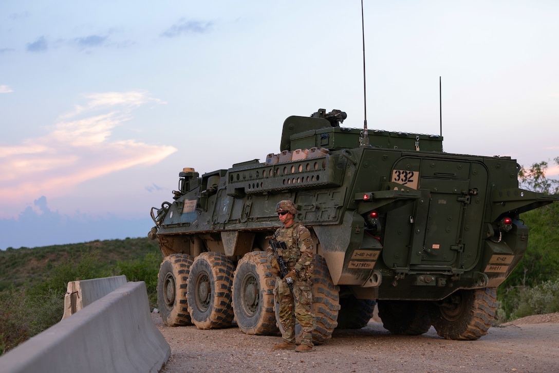 A soldier holding a rifle stands next to a vehicle parked on gravel and looks into the distance; green vegetation is in the background.