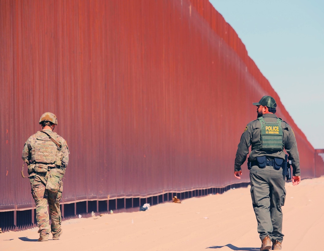 A soldier in combat gear and a Border Patrol agent wearing a green uniform walk on dirt next to a tall, rust-colored wall.