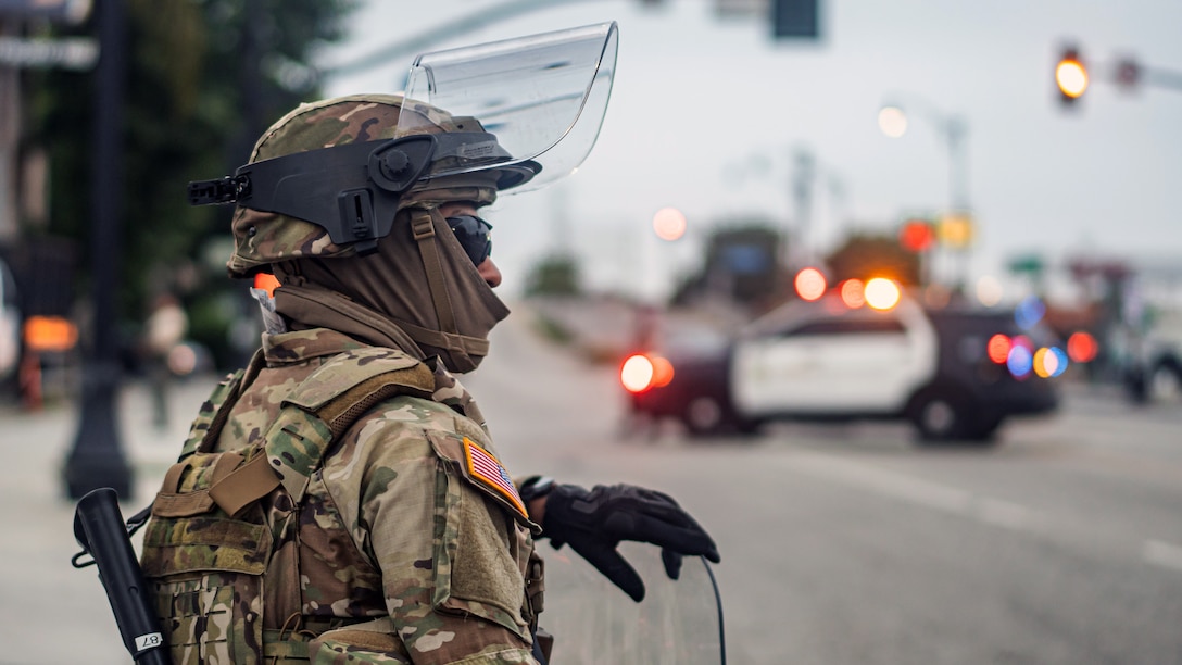 A soldier in combat gear and wearing black gloves stands near a road and a law enforcement vehicle with its lights on.