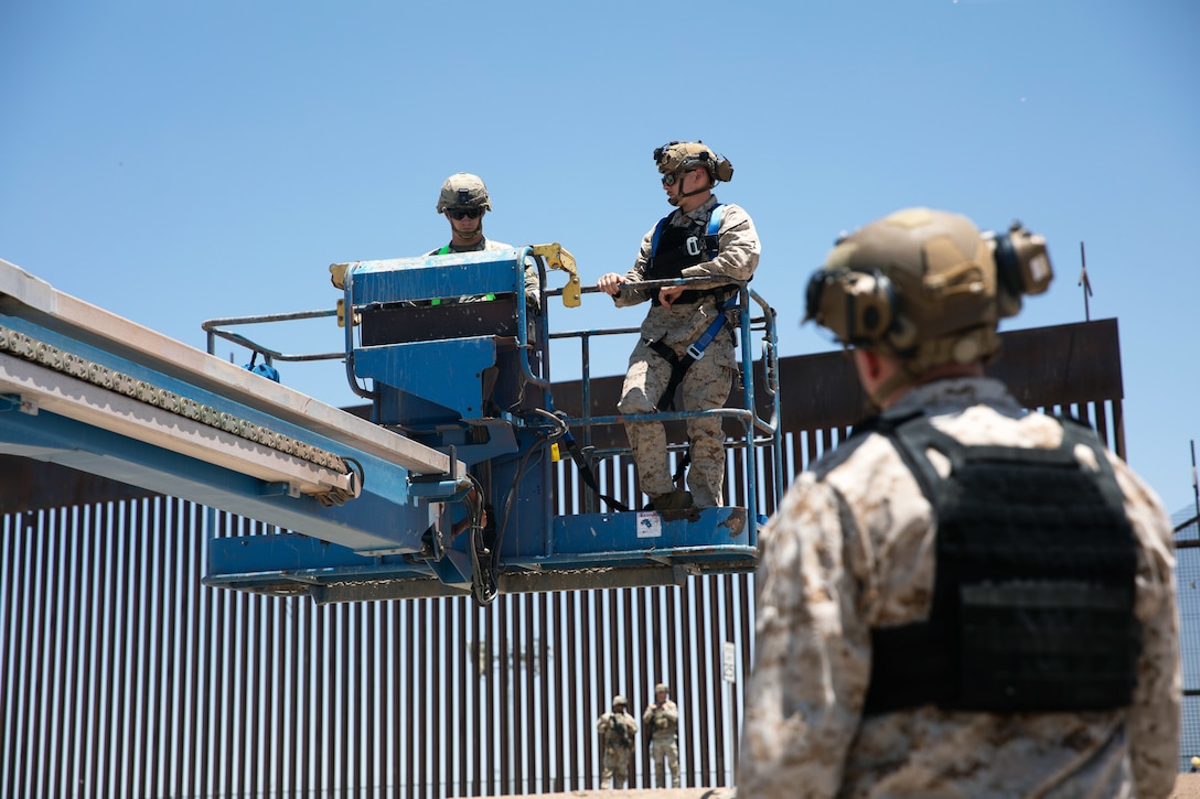 Two Marines watch a soldier operating a mobile elevated work platform near a barrier; two other troops stand in the background.