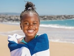 Young girl wrapped in blue and white towel stands on a beach.