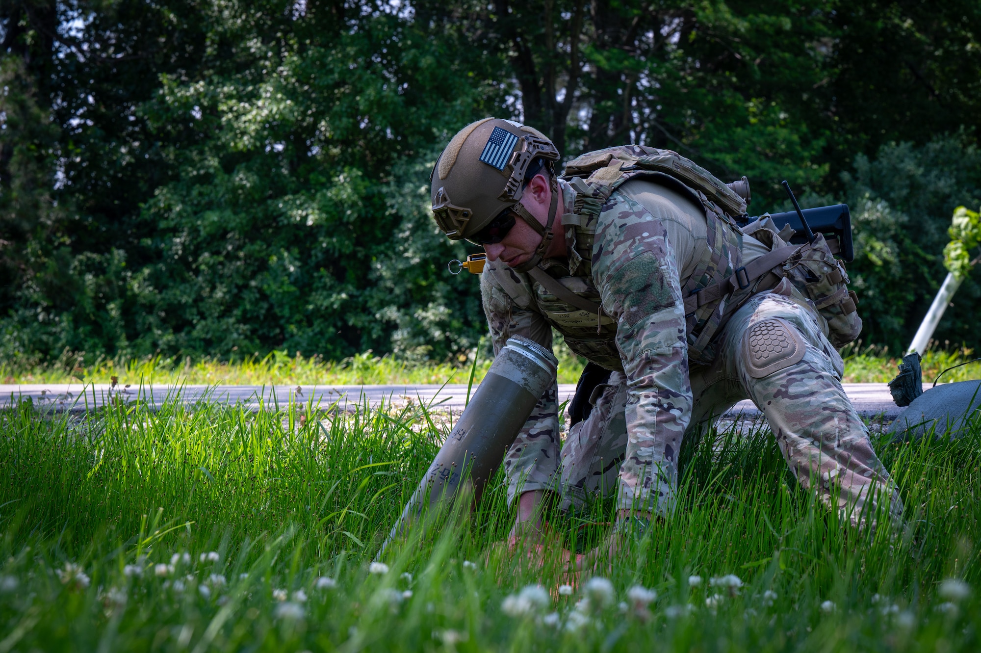 Staff Sgt Cox kneeling in the green grass because he has located a UXO. He is planting a marker near this location to warn others.