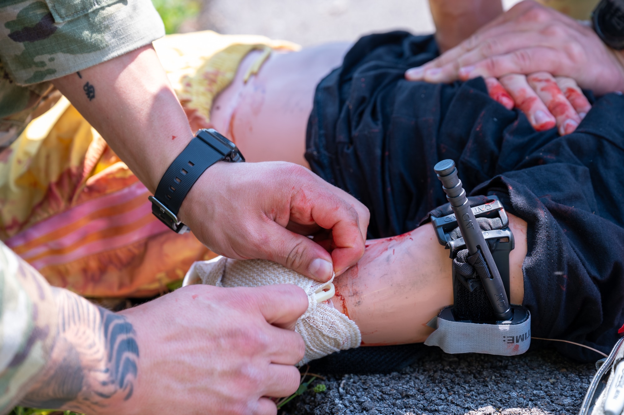 Close up shot of two Airmen's hands as they perform chest compressions and apply a tourniquet to a casualty mannequin laying outside on the asphalt.