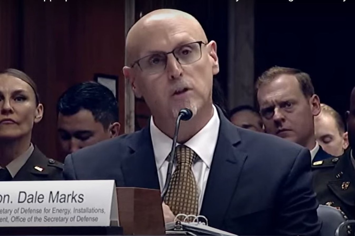 A man in civilian business attire is sitting at a table and speaking into a microphone.
