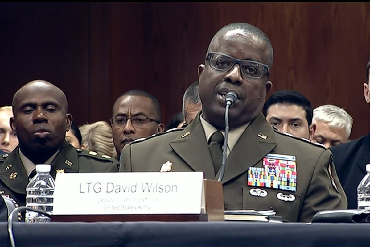 A man wearing a military dress uniform sits at a table while speaking into a microphone. Several men in military dress uniforms sit behind him.