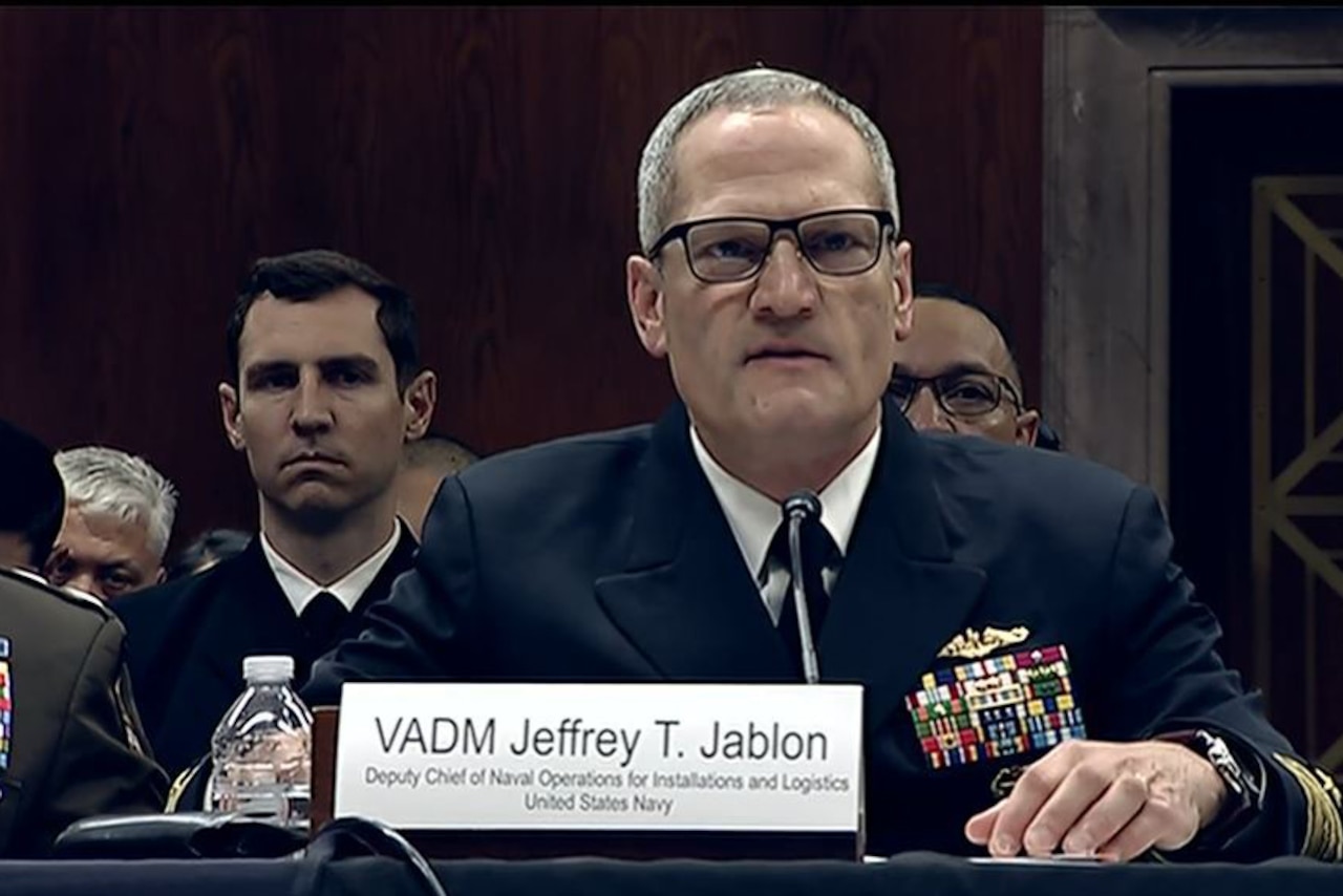 A man wearing a military uniform sits at a table while speaking into a microphone. Several men in military dress uniforms sit behind him.