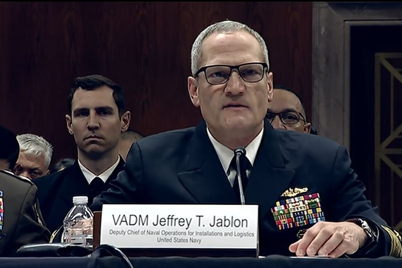 A man wearing a military uniform sits at a table while speaking into a microphone. Several men in military dress uniforms sit behind him.