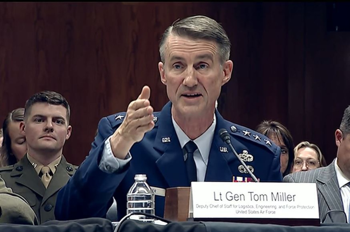 A man wearing a military dress uniform sits at a table while speaking into a microphone. Several men in military dress uniforms and business attire sit behind him.
