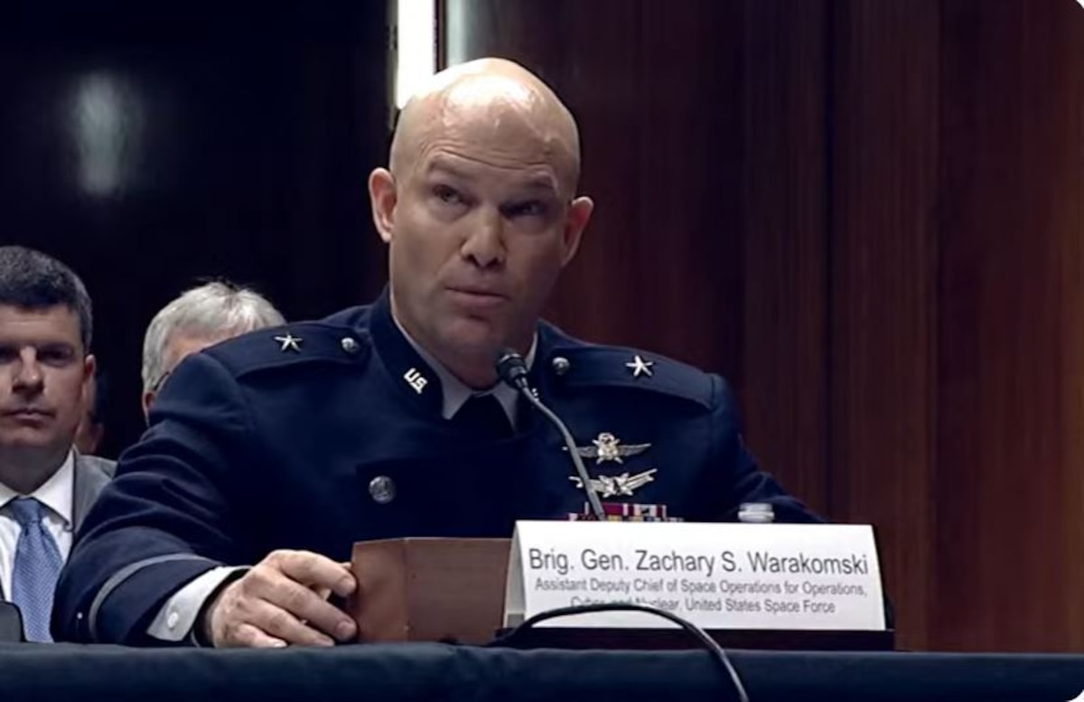A man wearing a military dress uniform sits at a table while speaking into a microphone. A man in business attire sits behind him.