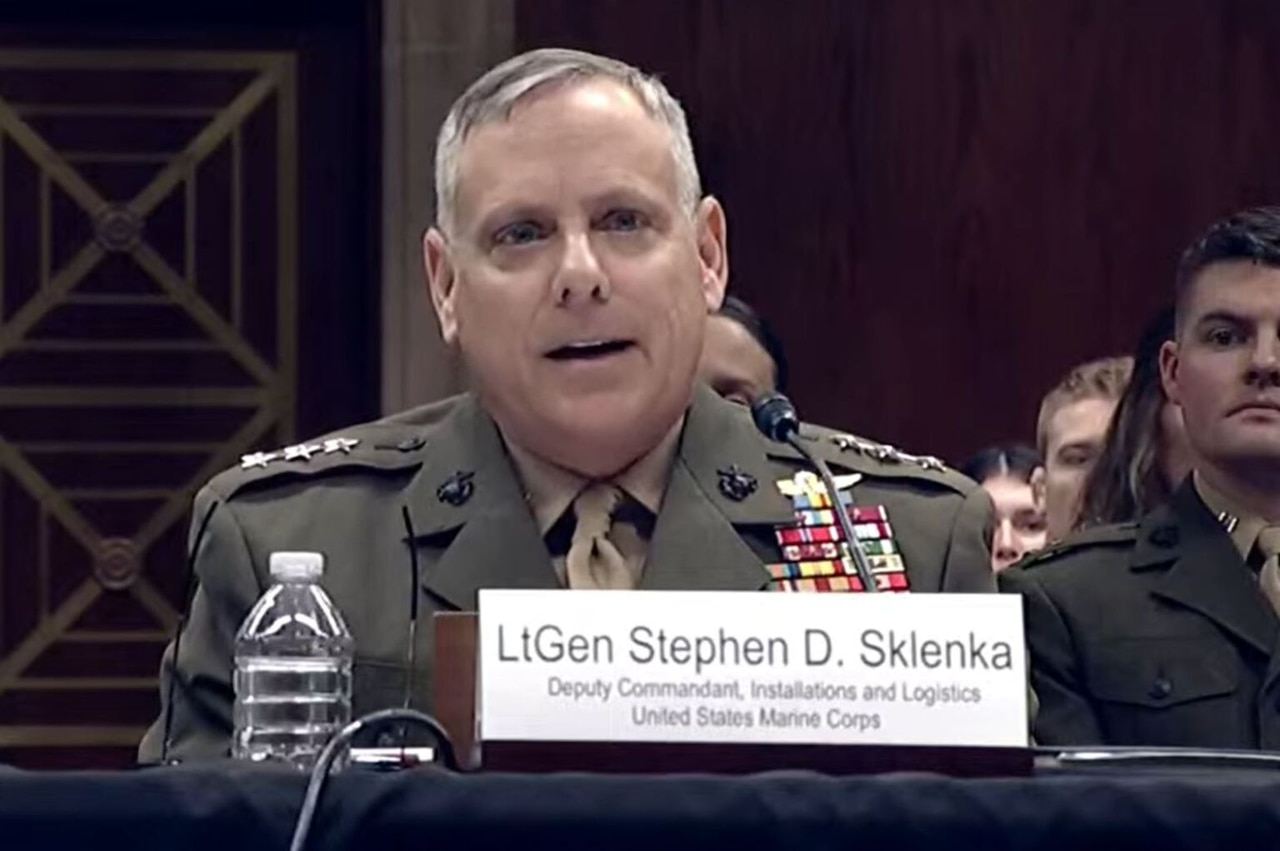 A man wearing a military dress uniform sits at a table while speaking into a microphone. Another man, wearing a military dress uniform, sits behind him.