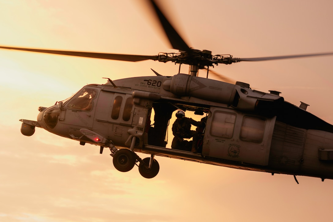 A sailor sits in a helicopter that is flying against a golden sky.