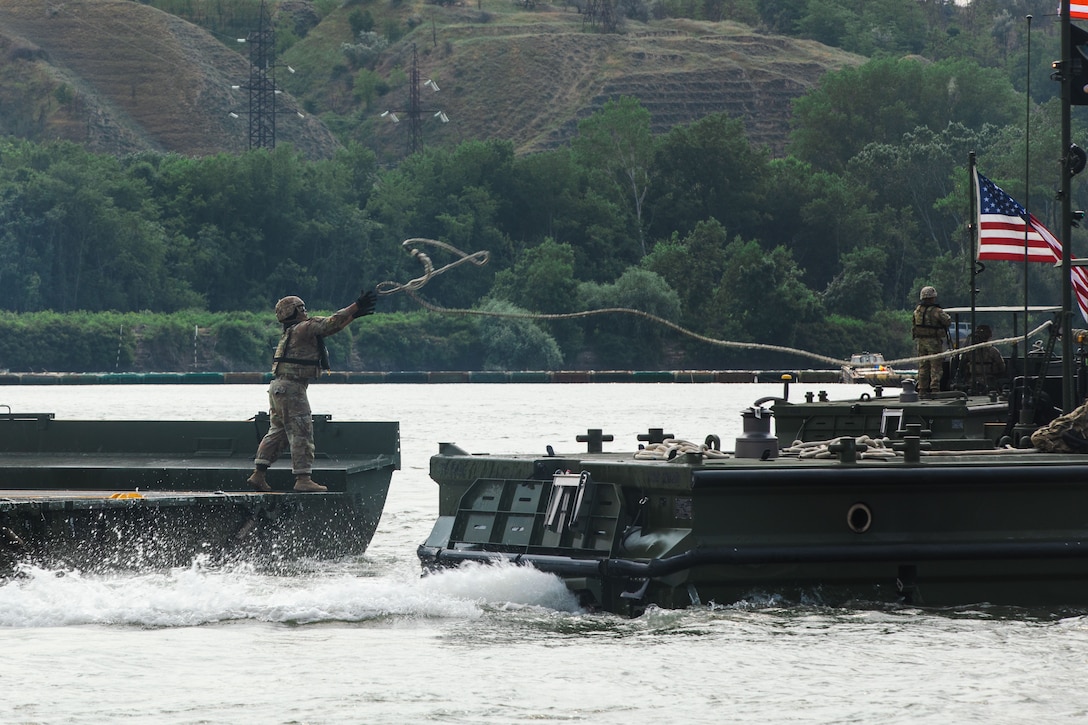 A soldier on a boat catches a rope thrown from another boat while sailing on a river.