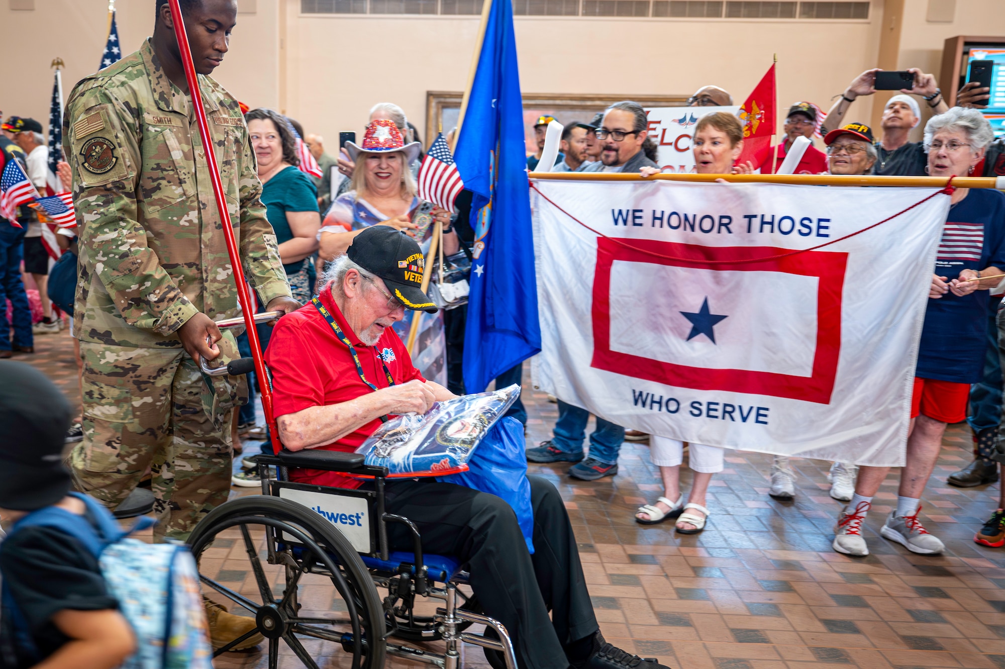 A service member pushes a man in a wheelchair through a crowd