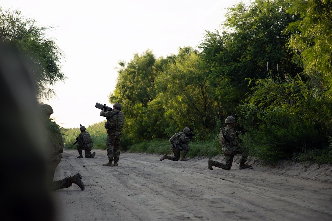 A soldier aims an anti-drone device toward the sky as others kneel on a dirt road with green vegetation on each side.