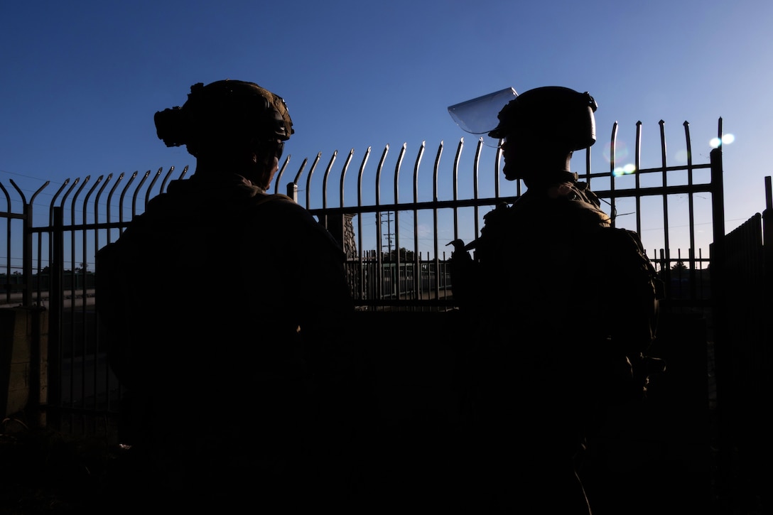 Two Marines wearing combat gear face a tall security gate with a blue sky in the background.