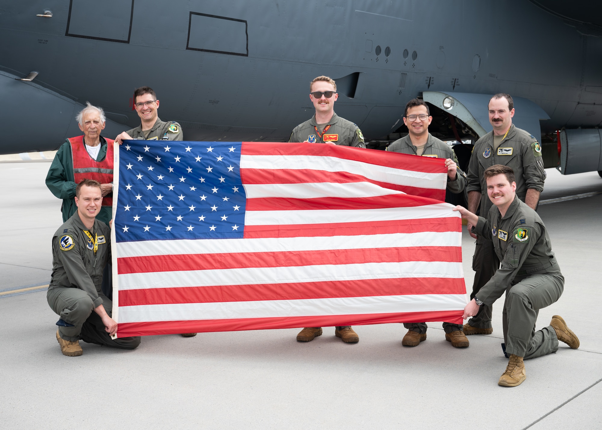 Gary Lee poses for a group picture with a 69th Bomb Squadron aircrew. The aircrew is holding open a flag of the USA.