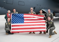 Gary Lee poses for a group picture with a 69th Bomb Squadron aircrew. The aircrew is holding open a flag of the USA.