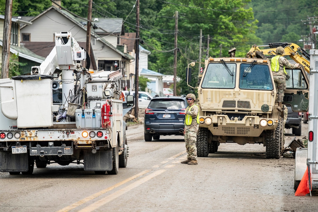 A guardsman works out of a utility truck as another guides traffic through trucks blocking a road.