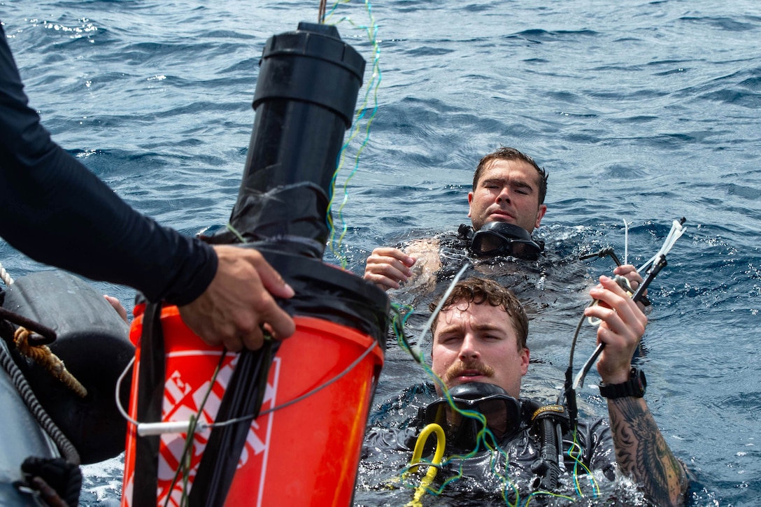 Two sailors treading water hold wires as a hand extends an explosive from an inflatable boat from the left.