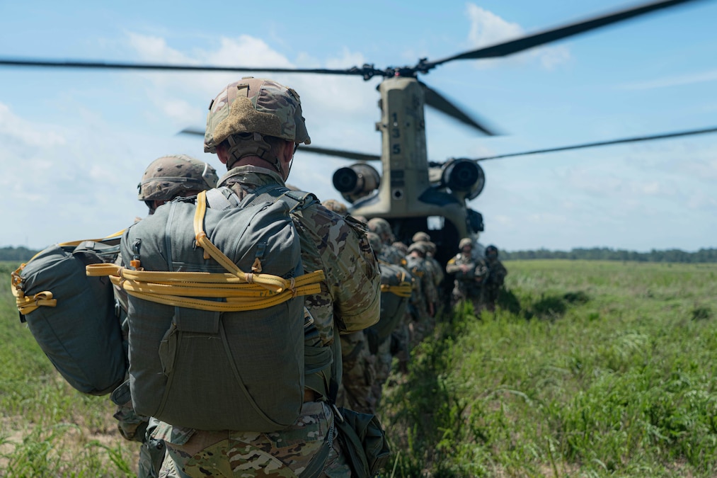 Multiple soldiers with parachute packs line up to enter a helicopter in a grassy field under a blue sky.
