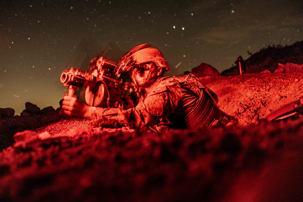 A Marine in red lighting aims a weapon while on the ground under the night sky.