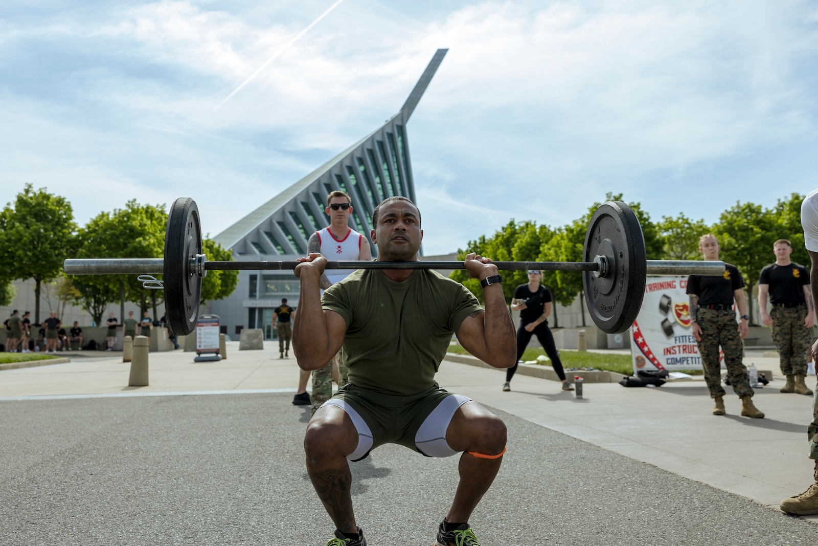 Marine in green physical training uniform performs a front squat with barbell and plates in front of the Marine Corps Musuem with multiple uniformed Marines in the background.